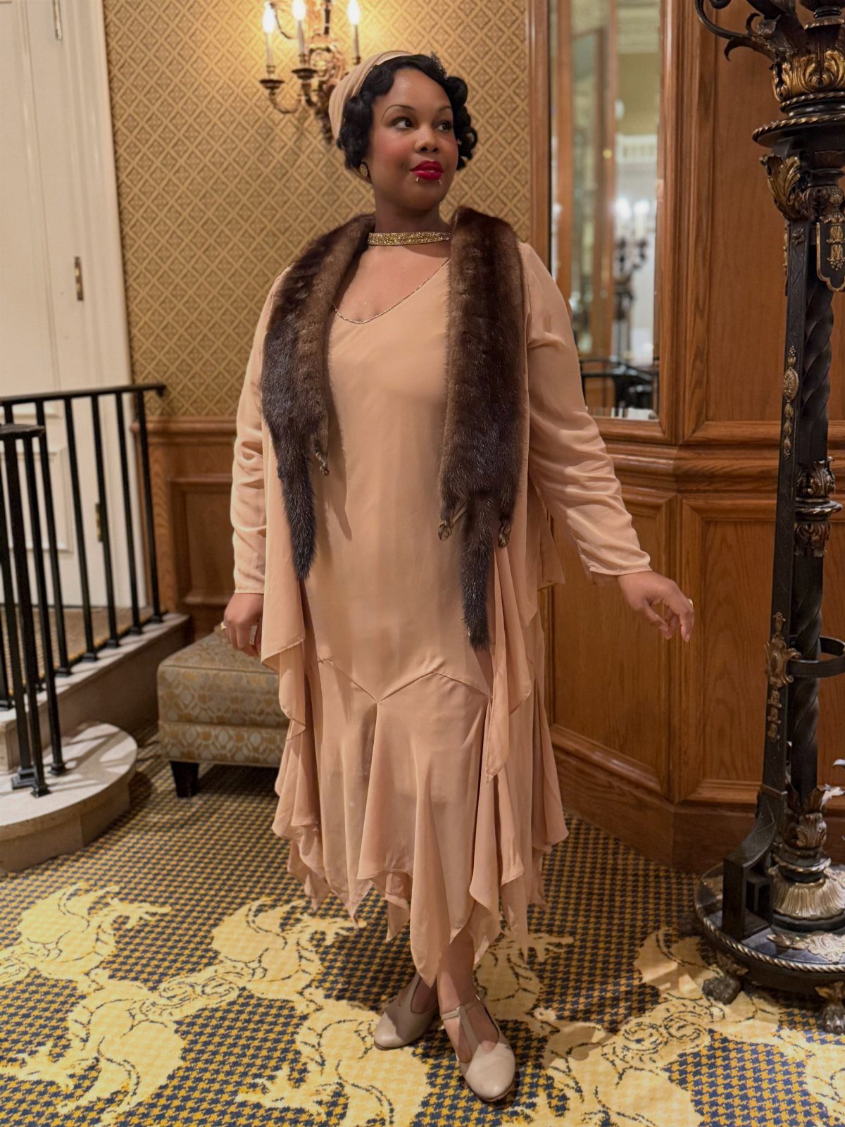 Woman in a peach 1920s dress and fur stole standing in an elegant room.