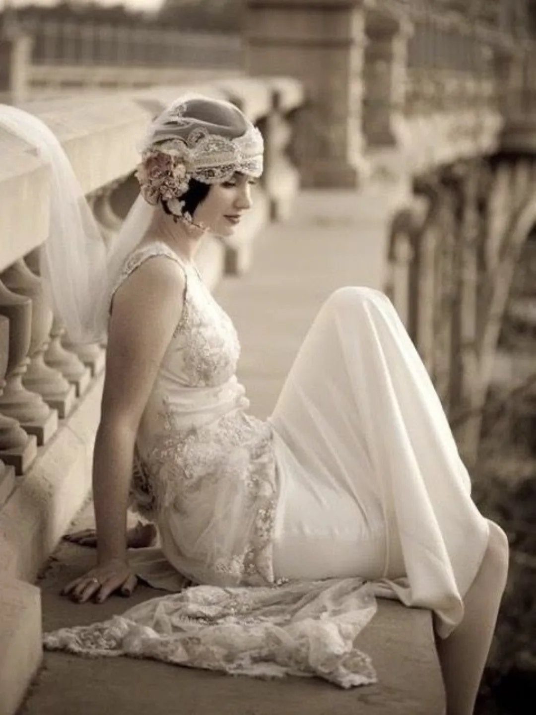 Woman wearing a vintage lace floor-length gown and beaded headpiece sitting on a stone ledge.