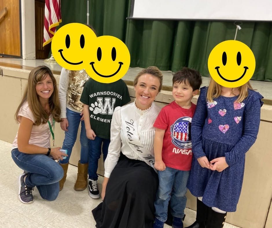 A woman wearing a white blouse and a Votes for Women sash posing with children.