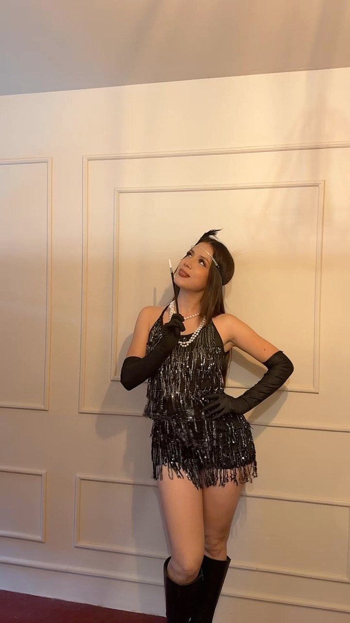 Woman in a short black fringed sequin dress and long black gloves posing against a wall.