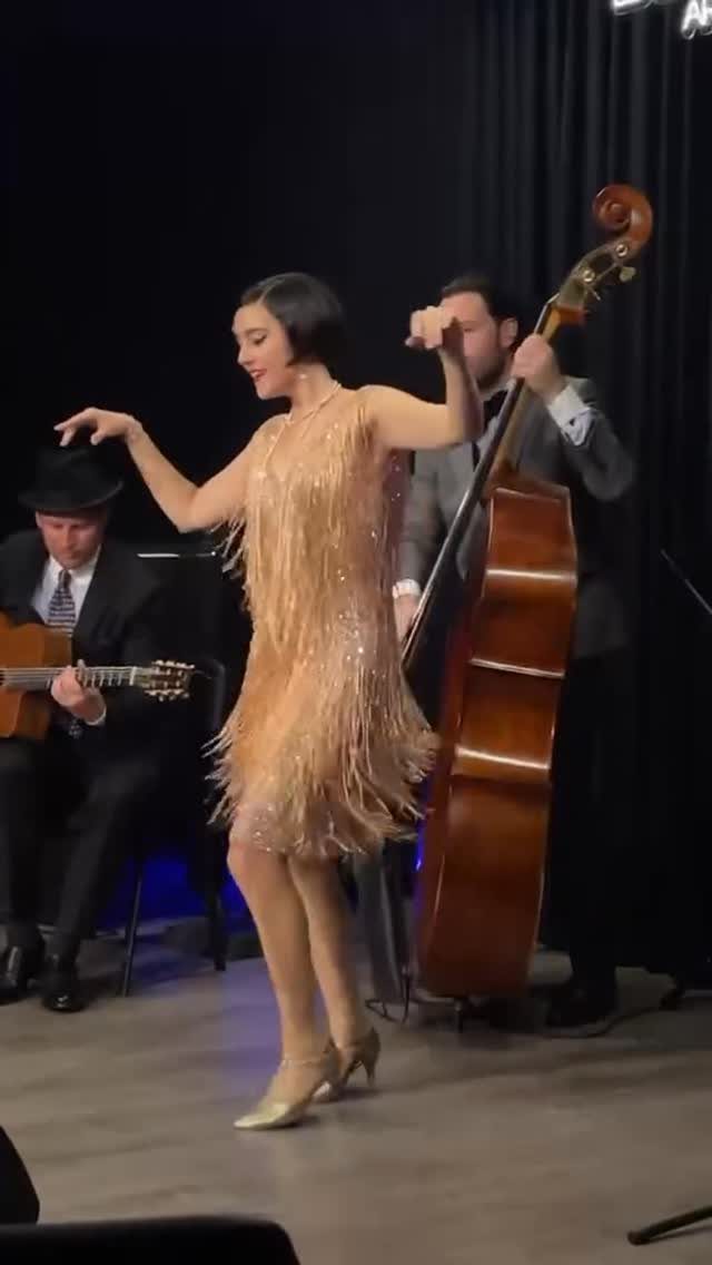 A woman wearing a gold fringed flapper dress dancing in front of musicians.