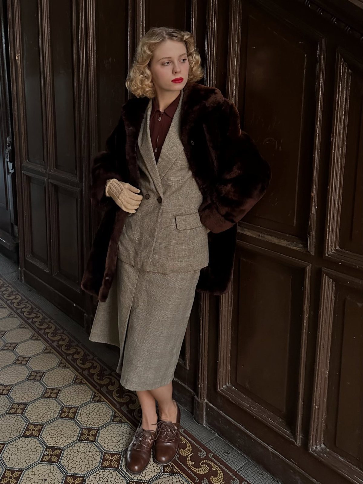 A woman in 1920s style tweed suit and fur coat standing by a wooden door.