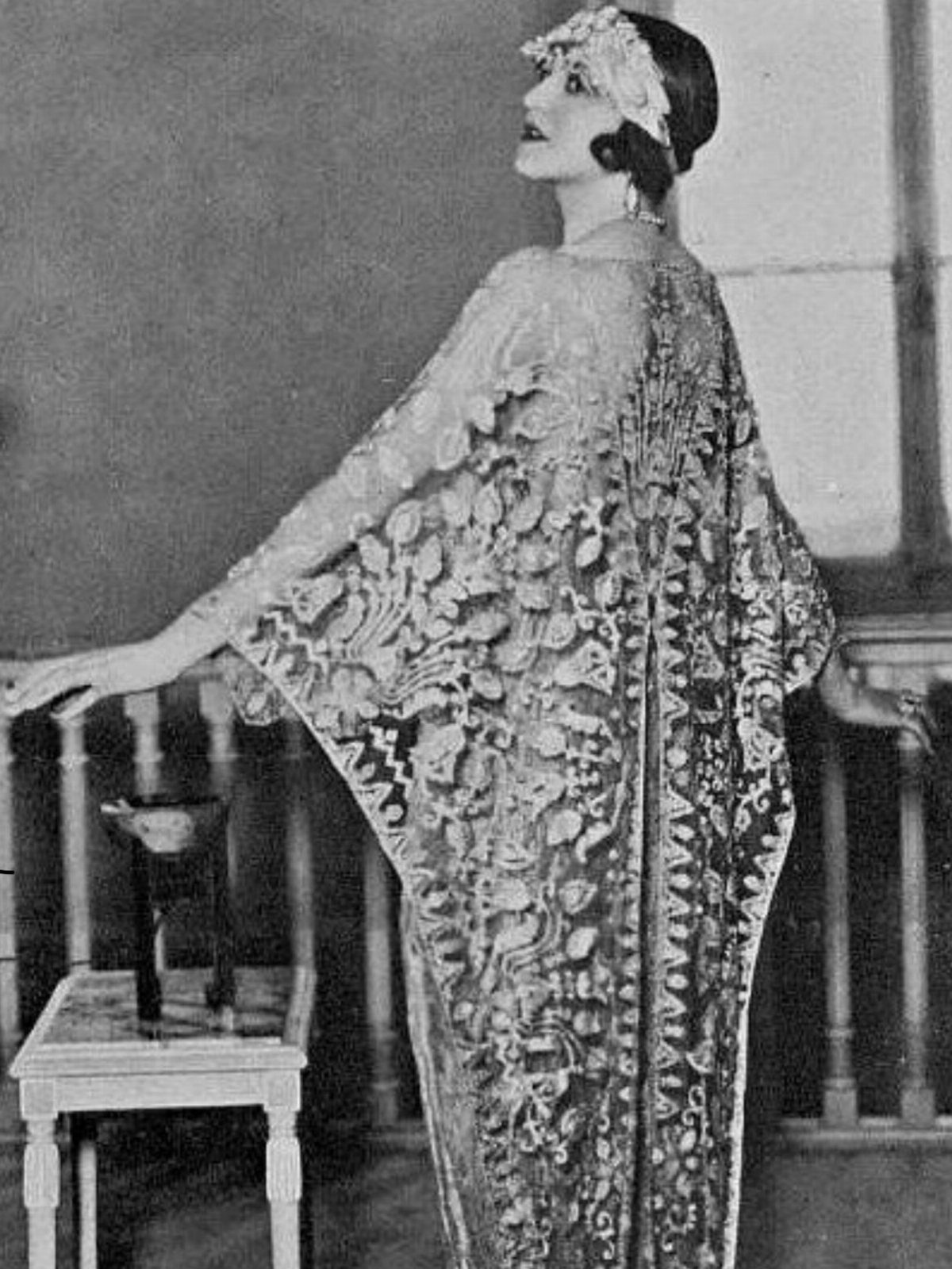 A woman in a patterned 1920s evening dress with a matching cape-like overlay stands by a railing.
