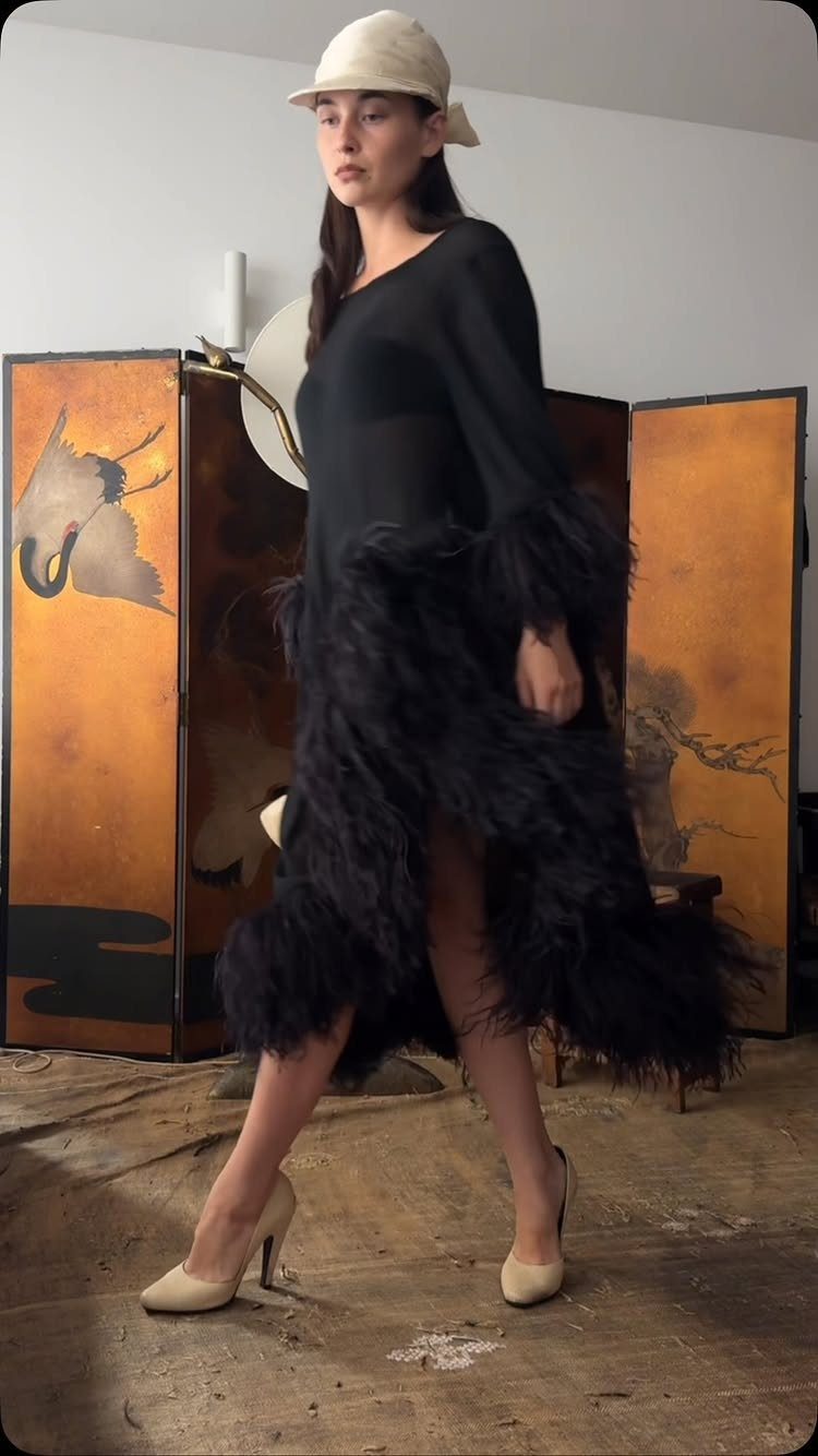 Woman in a black feathery evening dress and cream cloche hat posing before an orange screen.