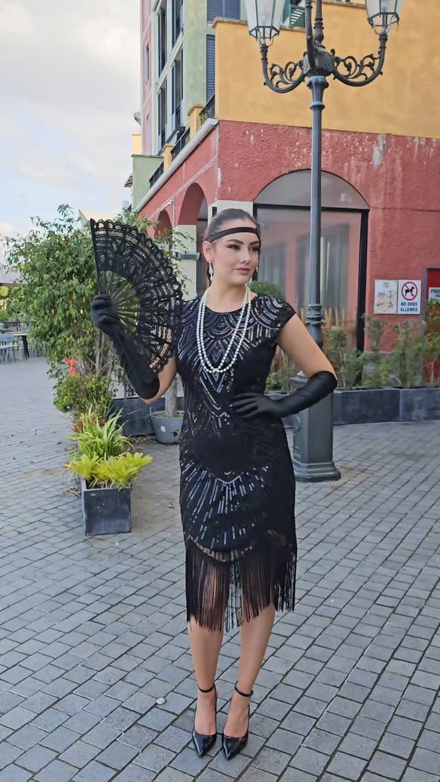 A woman in a black beaded flapper dress with fringe and long gloves holding a lace fan.