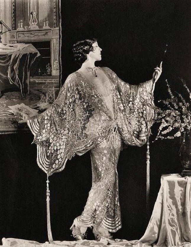 A woman in an embellished 1920s evening gown posing with a fan in a dark room.