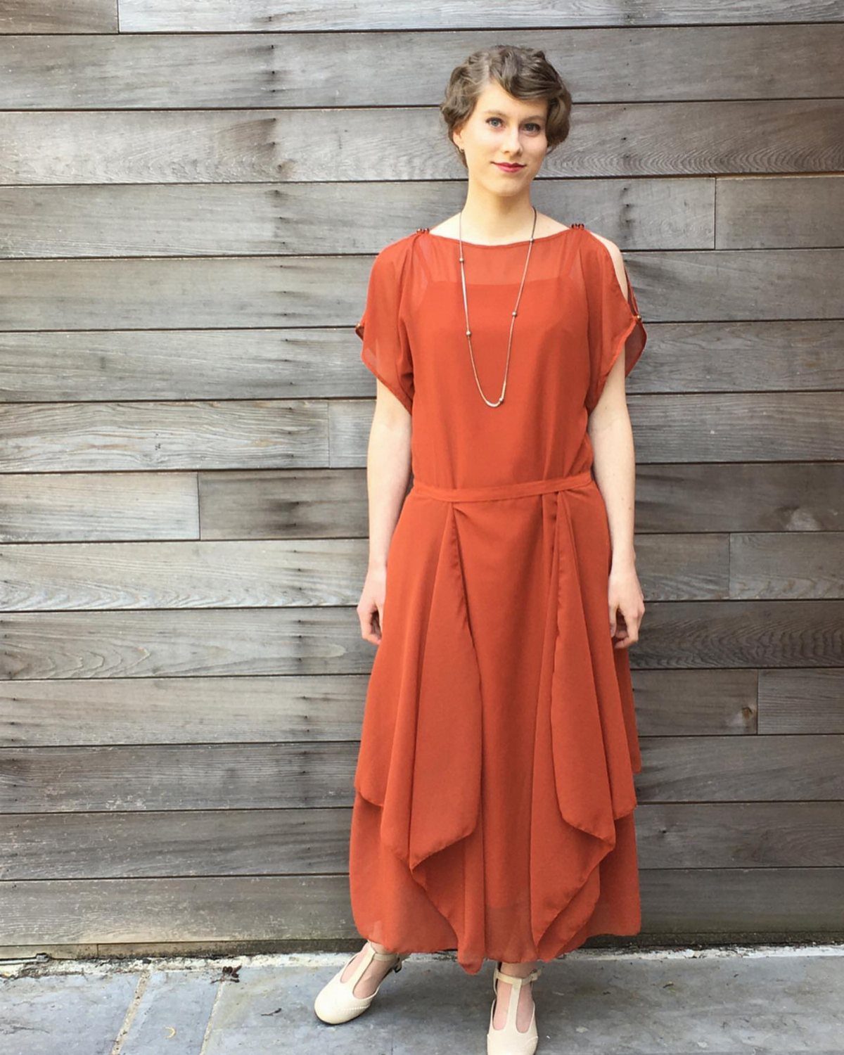 Woman in a burnt orange evening dress with a handkerchief hem standing against a wooden wall.