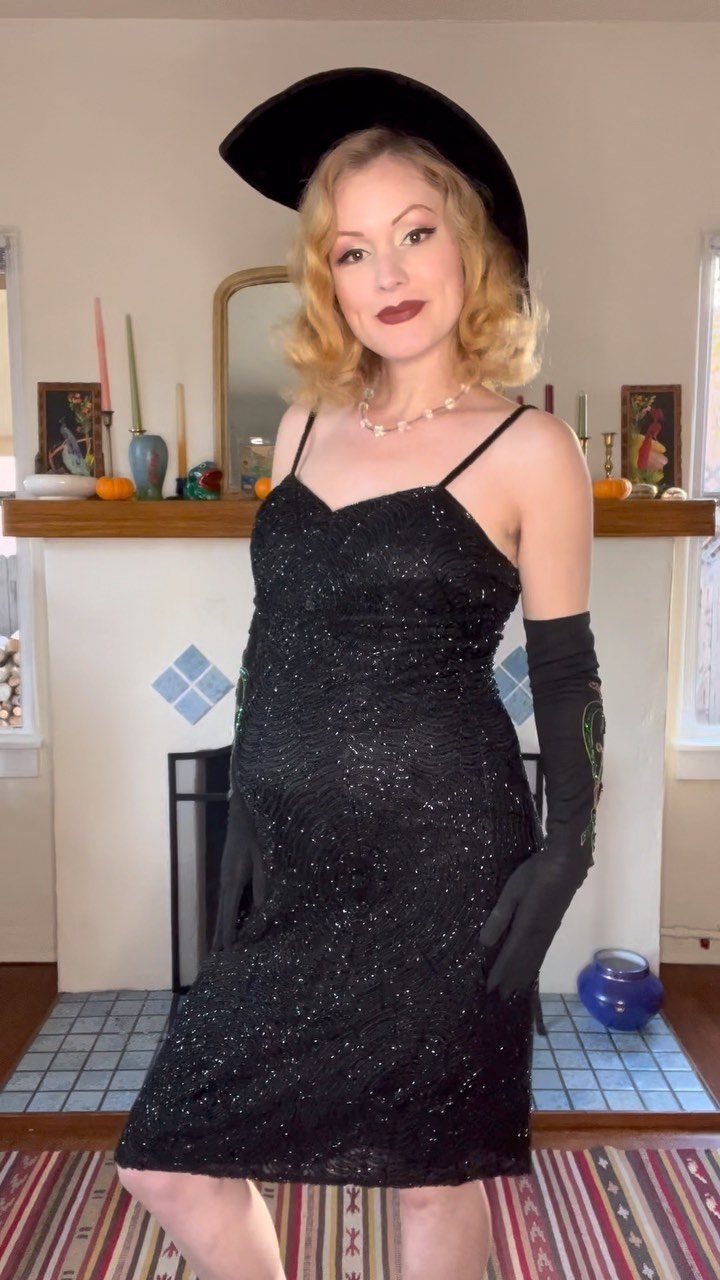 A woman in a black sequined dress, long gloves, and a wide-brimmed hat posing indoors.