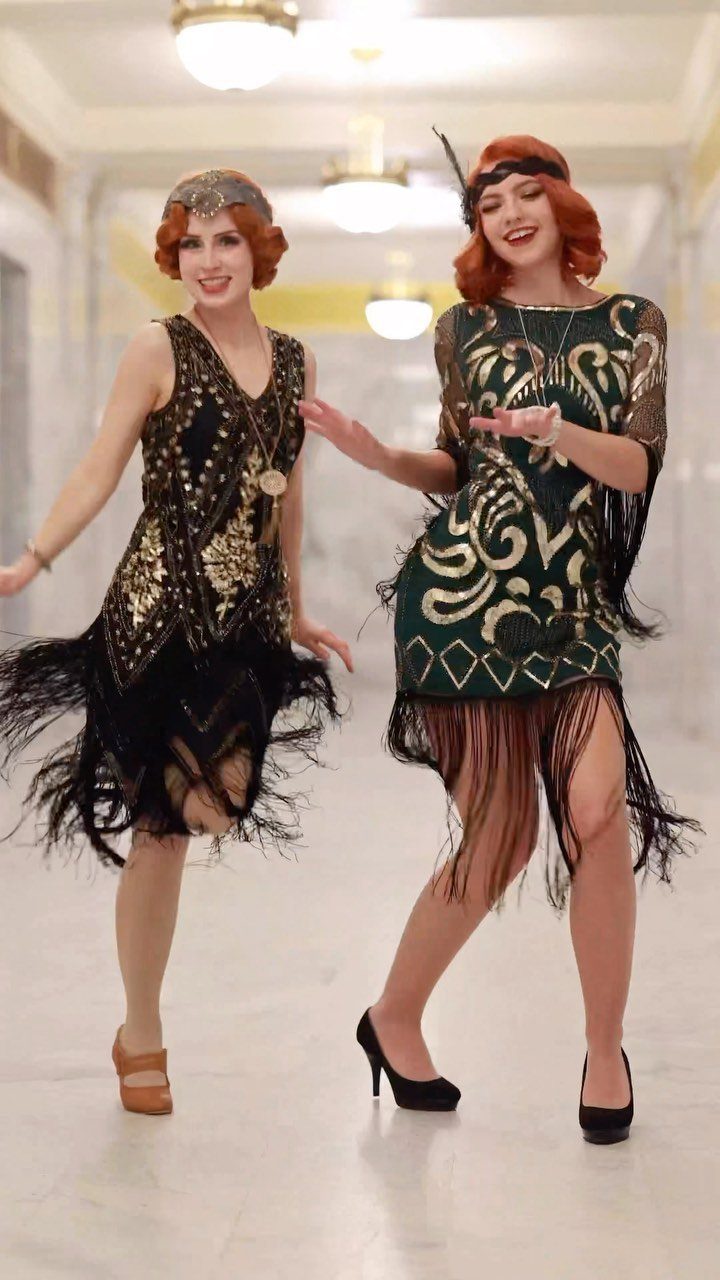 Two women smiling in sequined 1920s flapper dresses with fringe and headbands in a hallway.