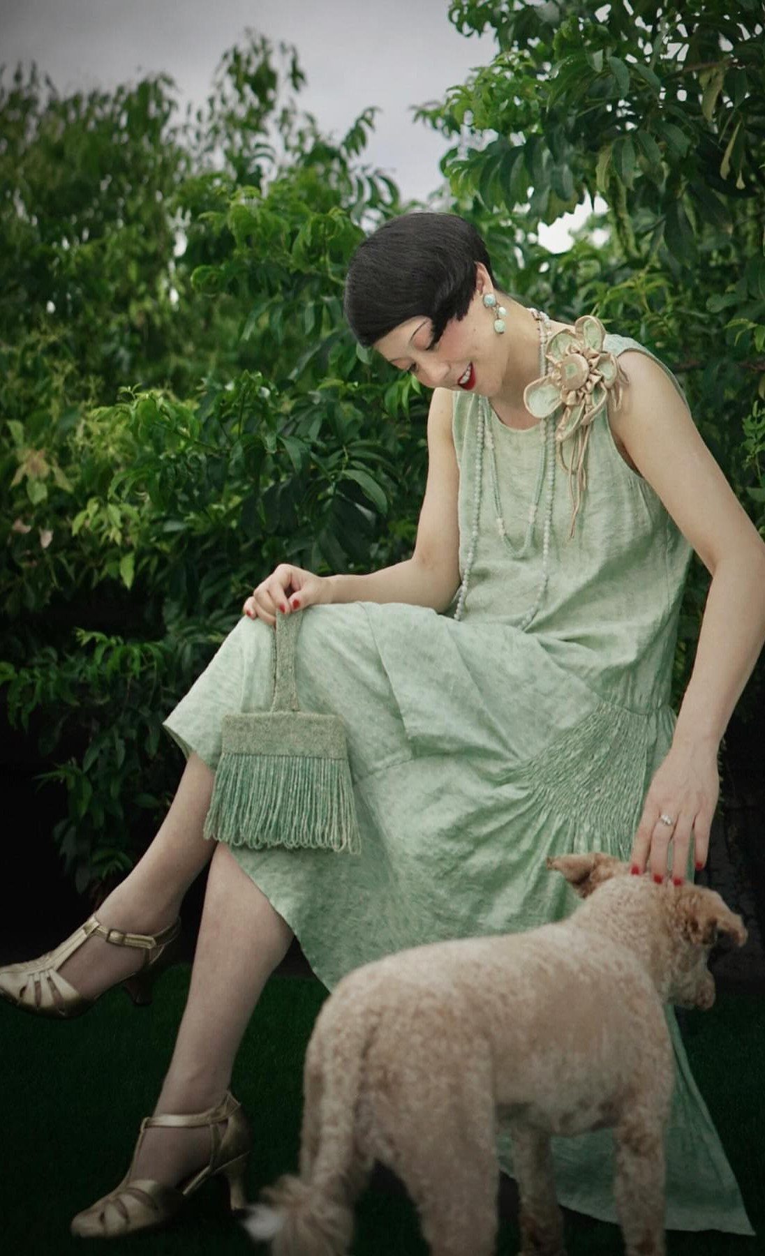 A woman in a light green sleeveless dress and gold heels sits in a garden.