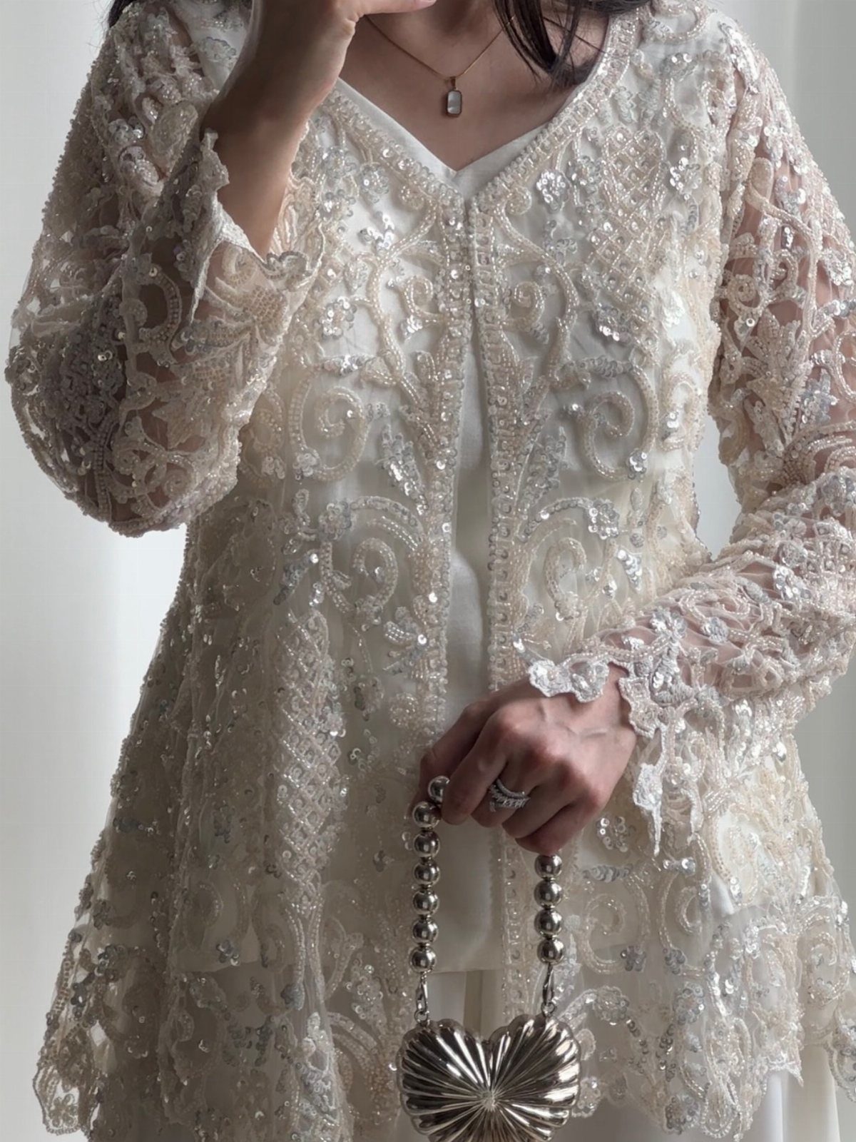 Cream lace jacket with intricate embroidery and sequins worn over a white dress with a silver purse.