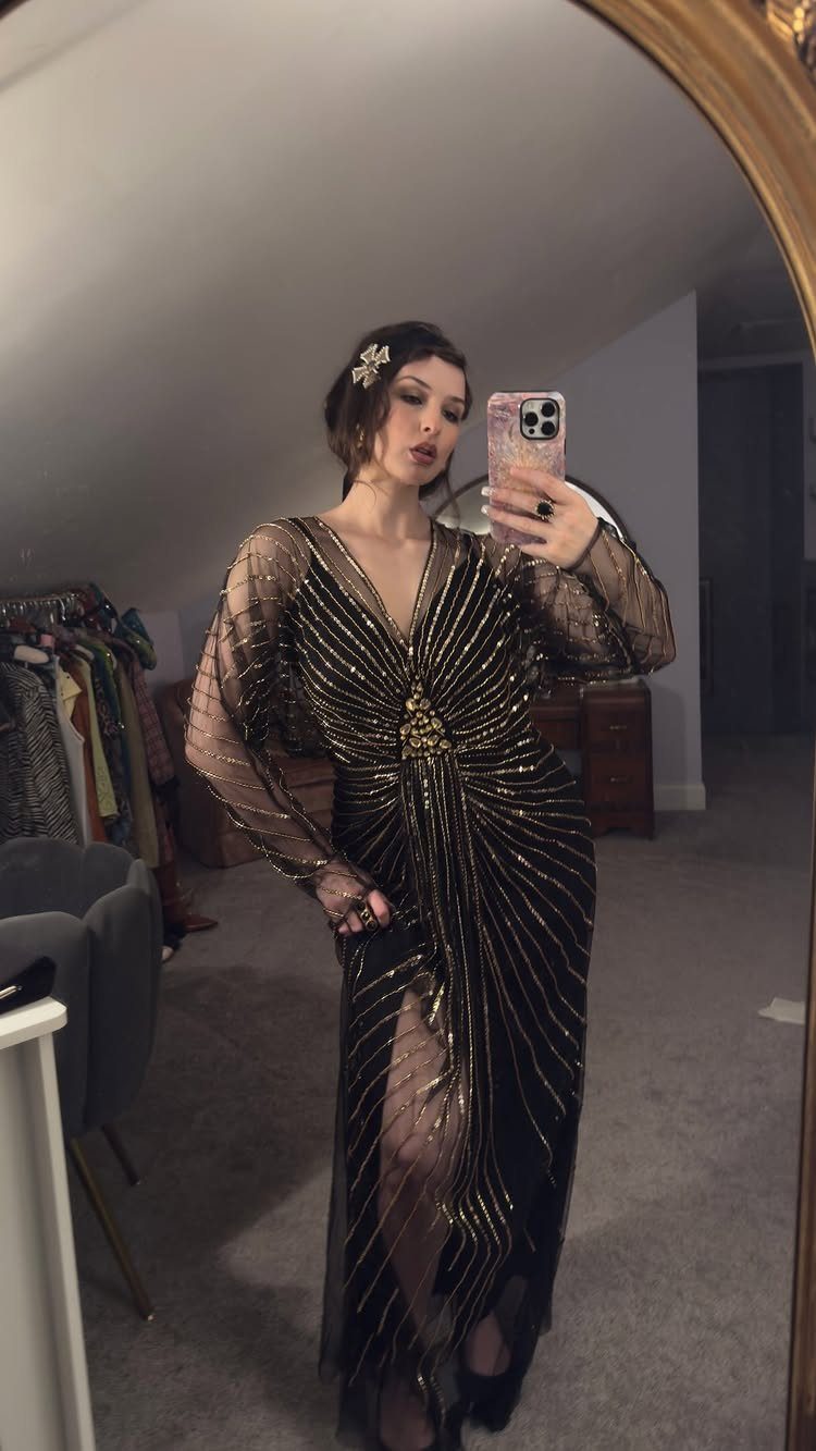 Woman in a black and gold sequined long gown posing for a mirror selfie.