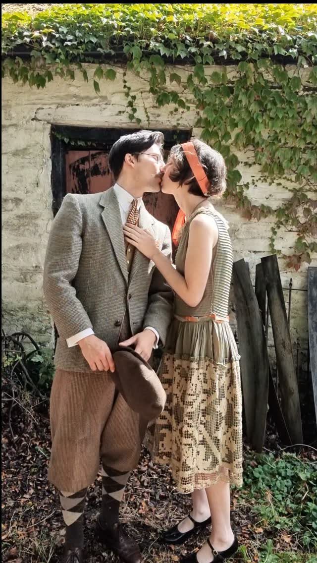 A man in vintage tweed clothing and a woman in a drop-waist dress kiss outdoors.