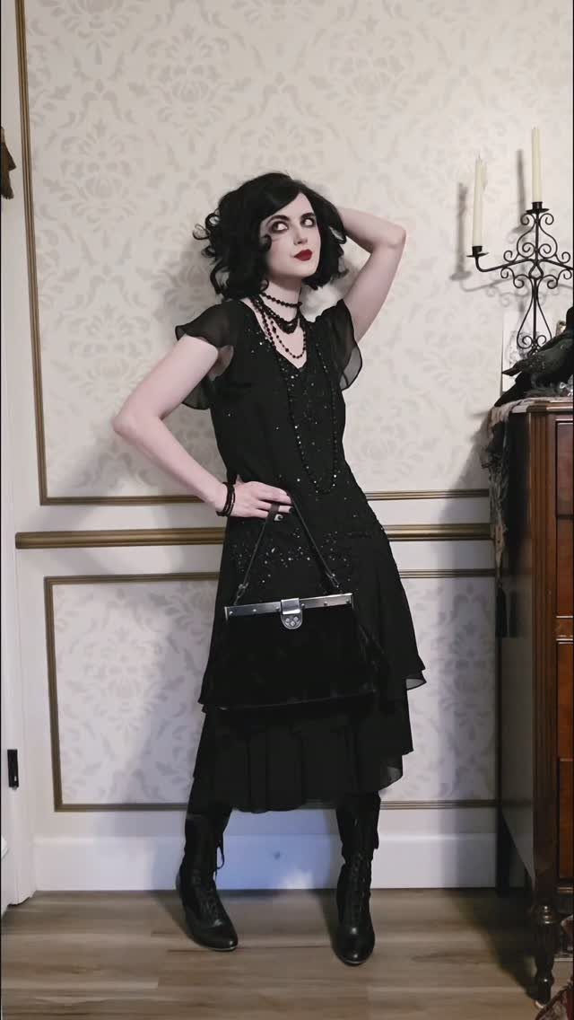 Woman in a black beaded drop waist dress posing with a black handbag.