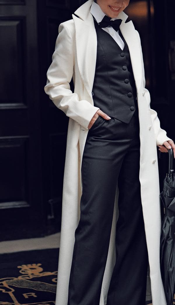 Person wearing a long white coat, black waistcoat, bowtie, and black trousers standing outdoors.
