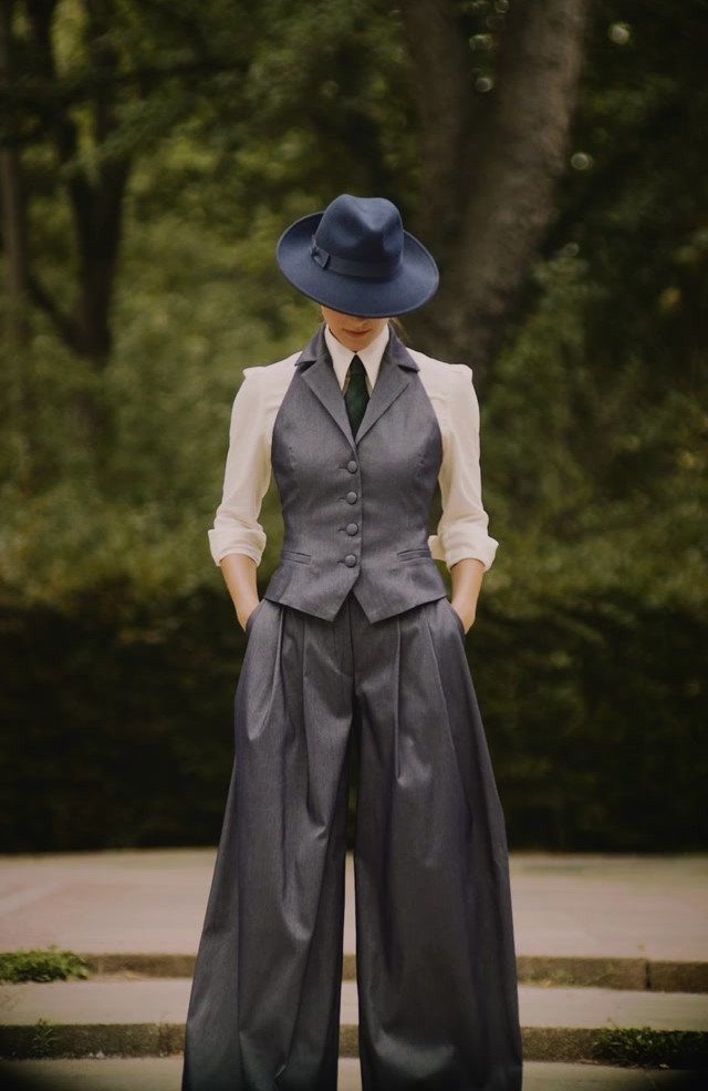 A woman wearing a blue hat, white blouse, grey waistcoat, and wide-leg trousers standing outdoors.