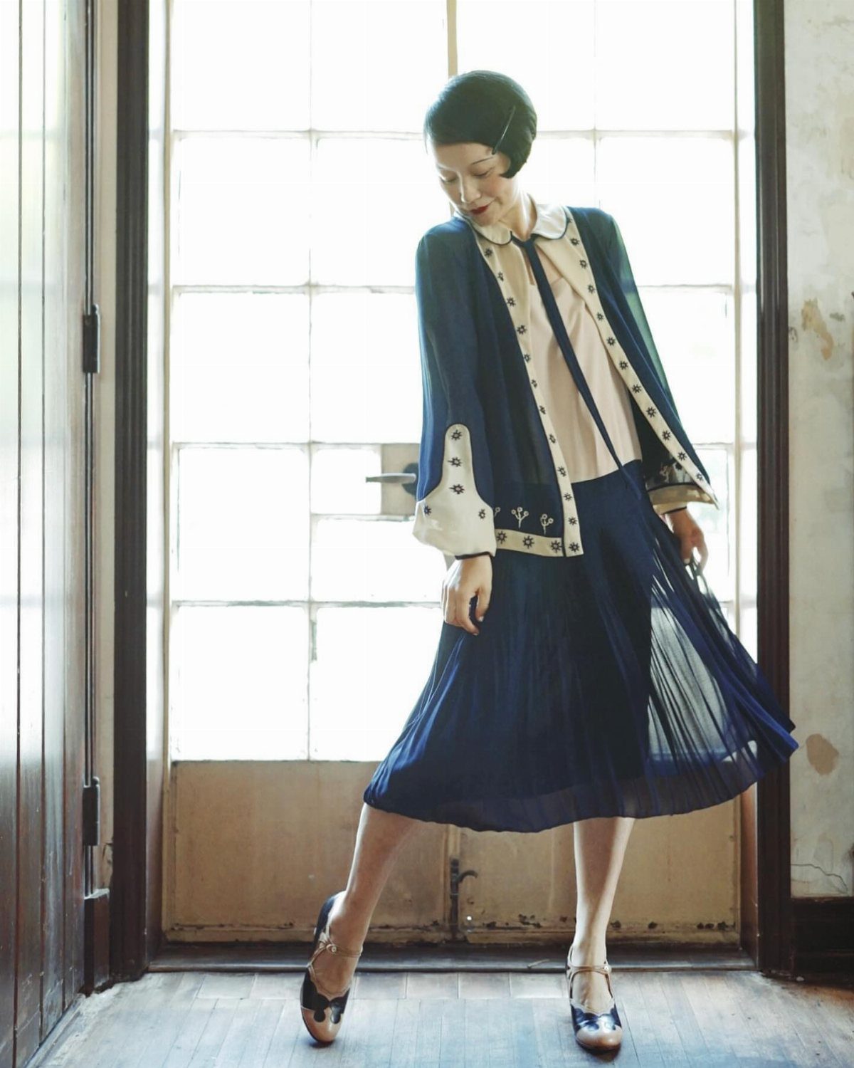 A woman posing in a blue 1920s flapper outfit with a pleated skirt and T-strap shoes.