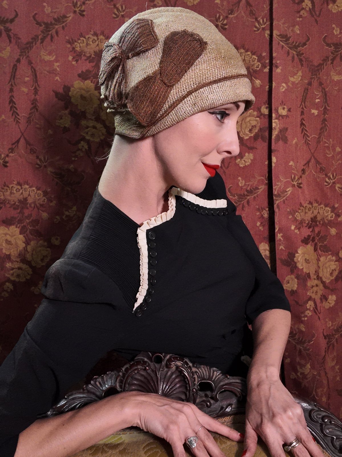 A woman wearing a patterned tan cloche hat and black top against a red floral background.