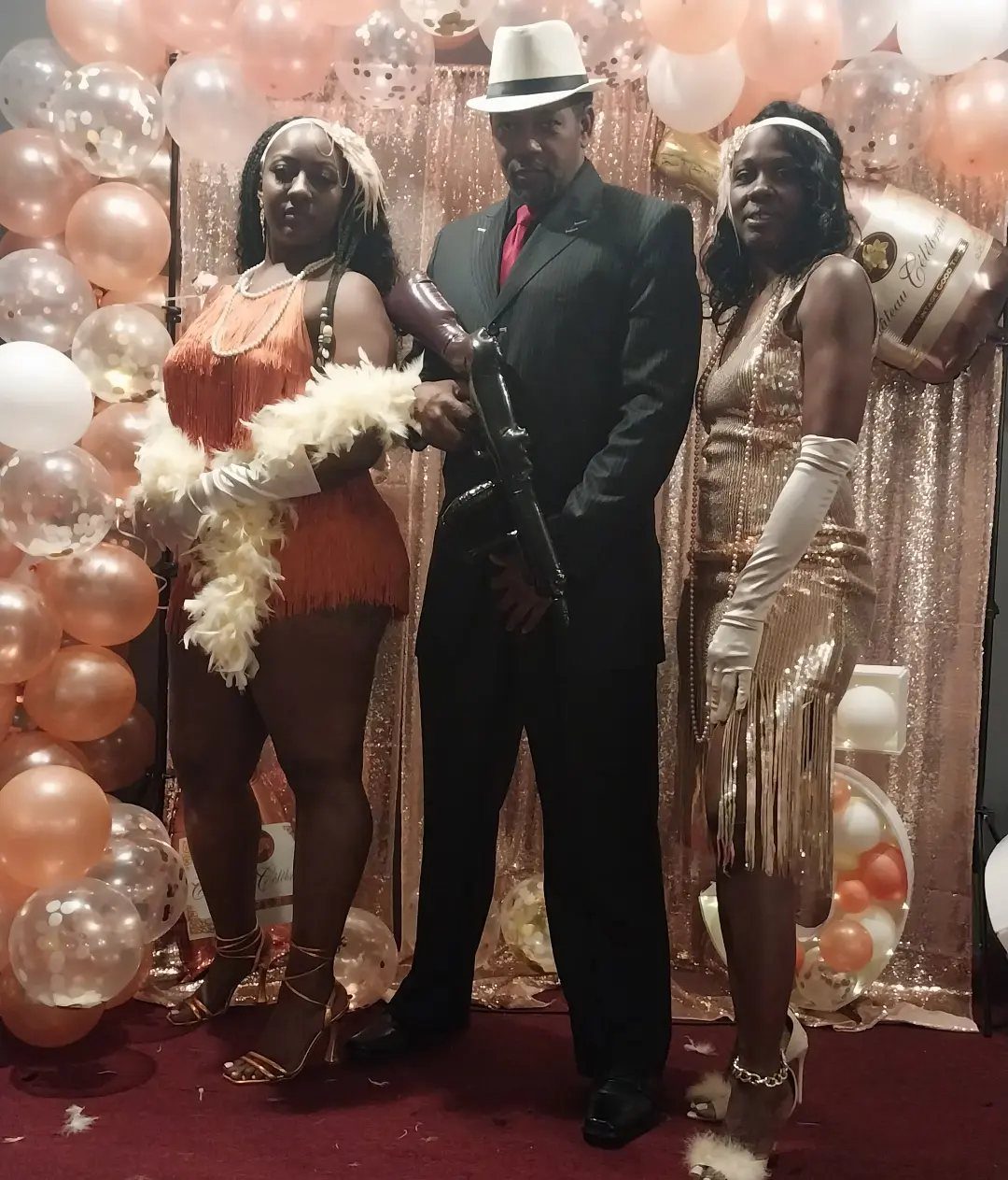 Three people dressed in 1920s party costumes posing in front of rose gold balloons.