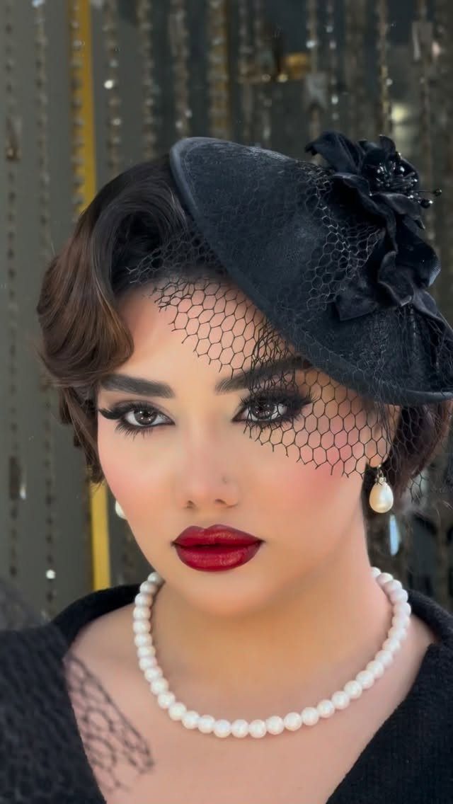 Woman with dark wavy hair wearing a black veiled hat, pearl necklace, and bold red lipstick.