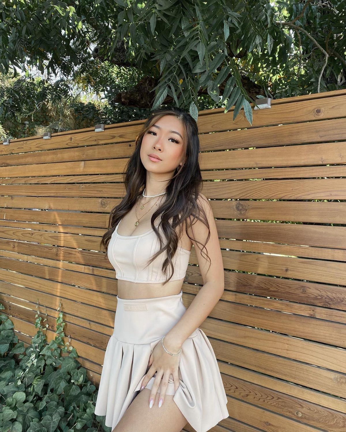 A woman in a cream two-piece crop top and skirt posing against a wooden fence.