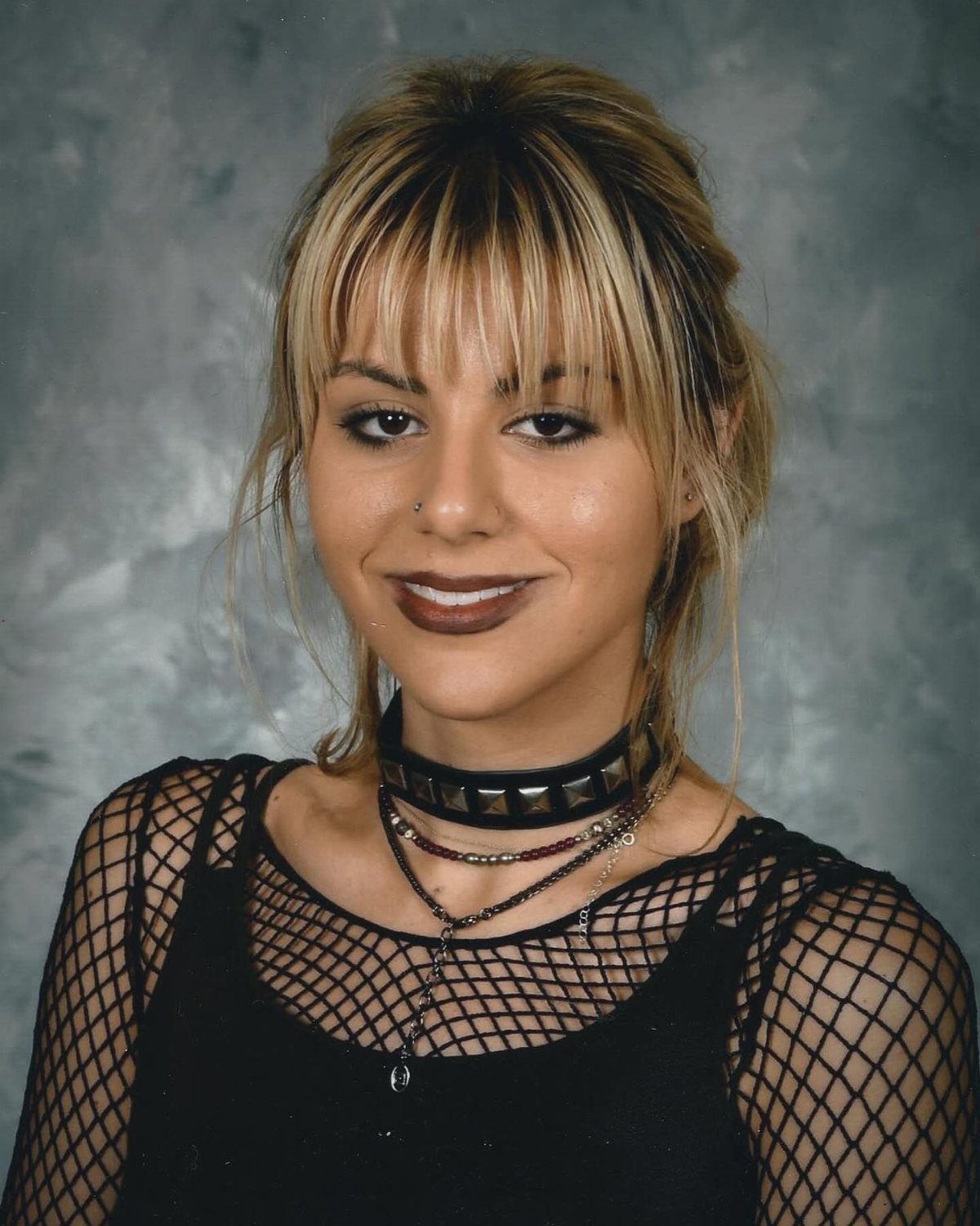A young woman with blonde hair wearing a black studded choker and fishnet top.