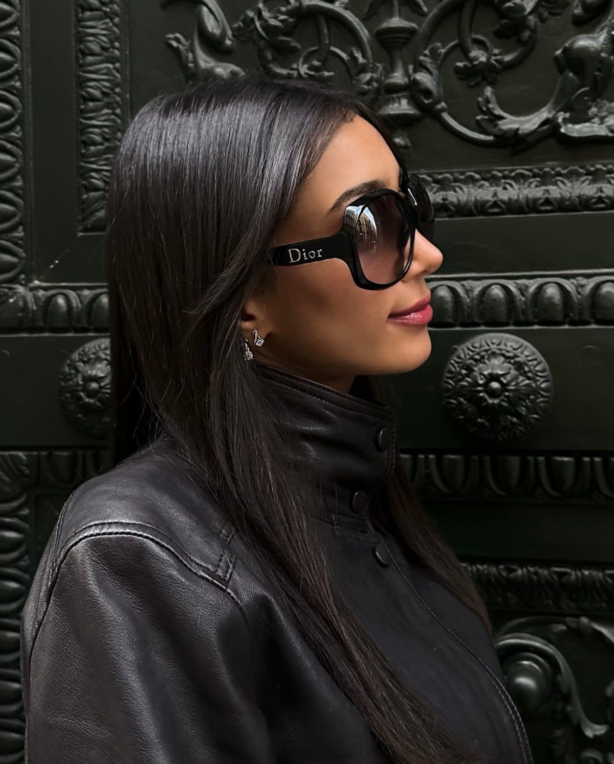 Woman with long dark hair wearing large black Dior sunglasses and a black leather jacket.