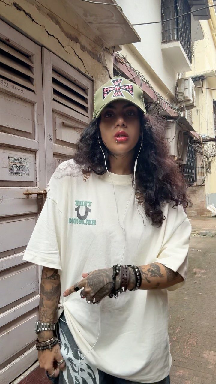 A woman wearing a graphic t-shirt, trucker hat, and multiple bracelets standing in an alley.