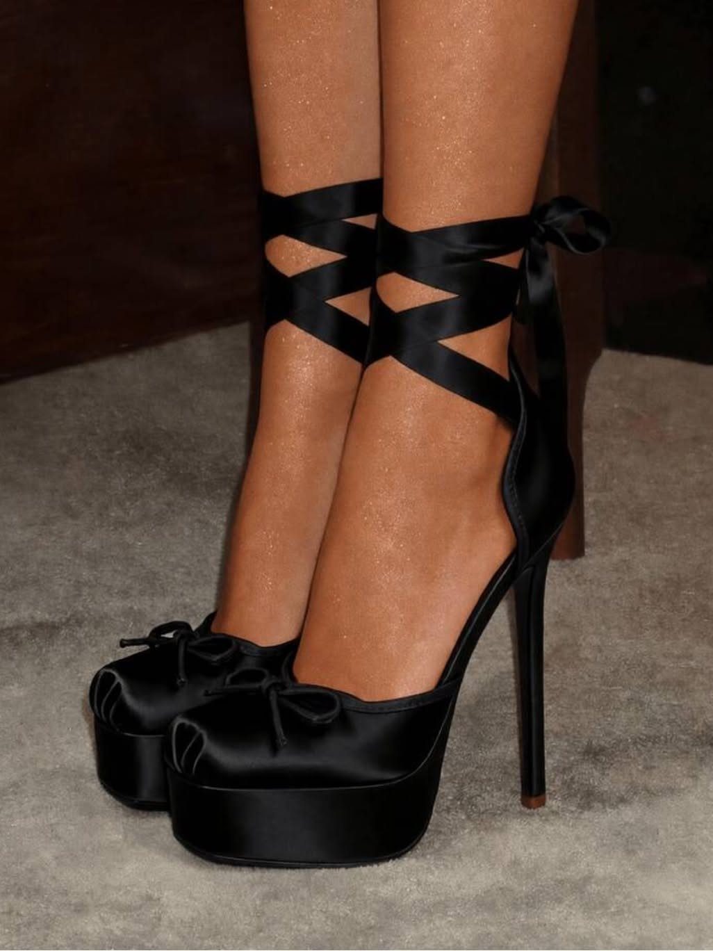 Black satin platform high heels with ribbon straps wrapped around the ankles on a carpeted floor.