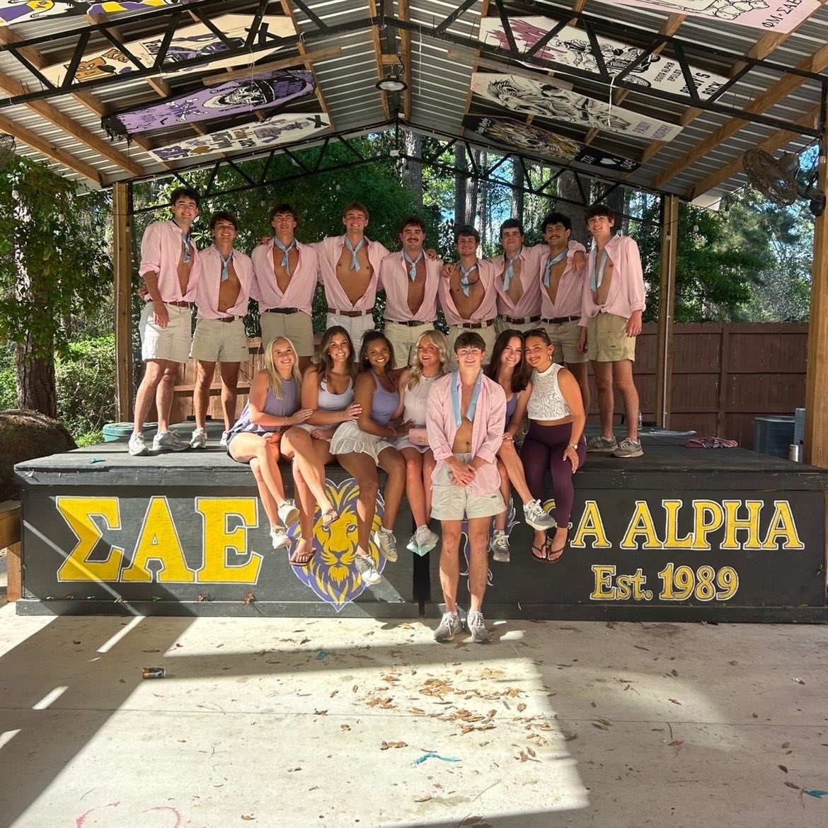 Young men in pink shirts and women in summer attire posing under a wooden pavilion.