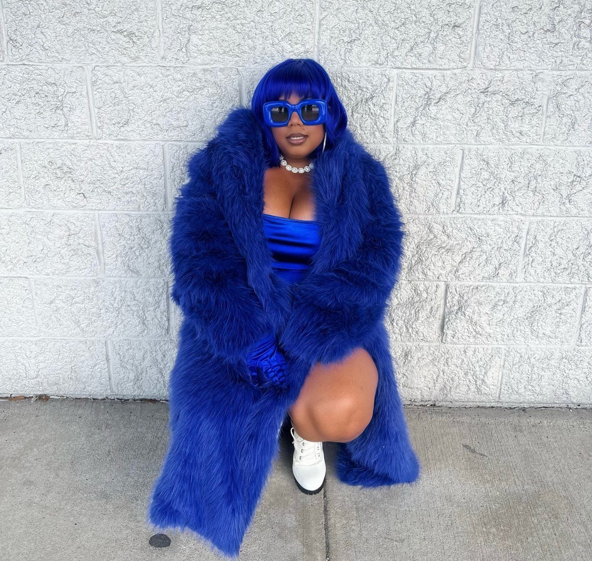 A person in a bright blue faux fur coat and blue satin outfit posing against a brick wall.