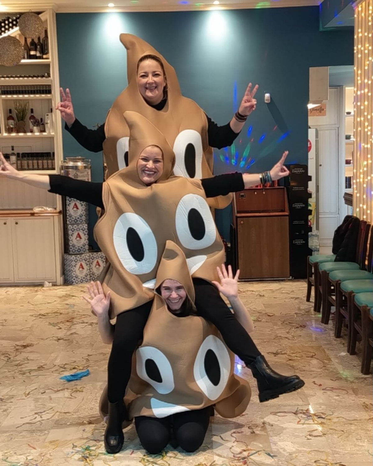 Three women posing in large, brown, plush poop emoji costumes in a room.