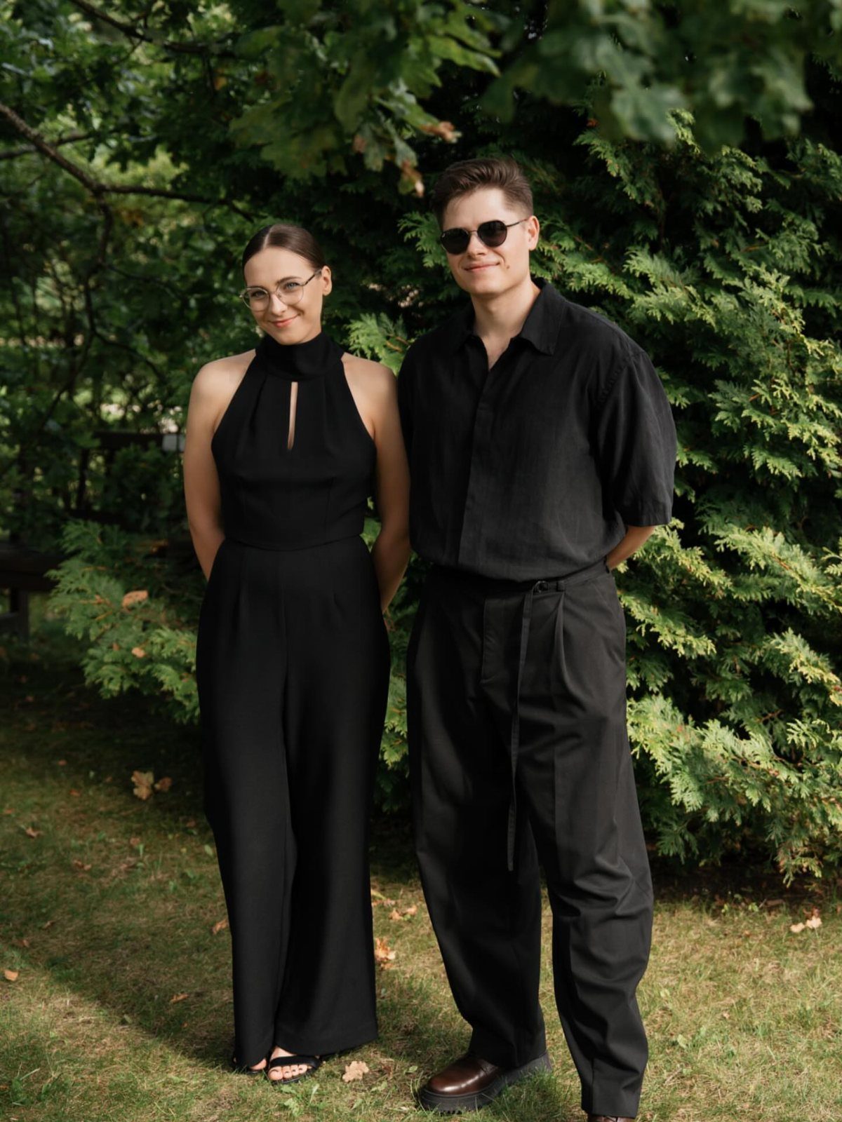 A woman in a black halter jumpsuit and a man in black clothing standing before green bushes.