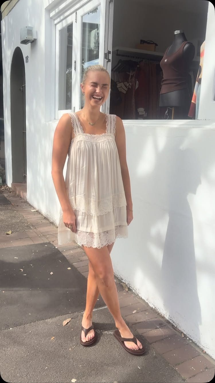 A smiling woman wearing a cream lace babydoll dress and flip-flops standing outdoors.
