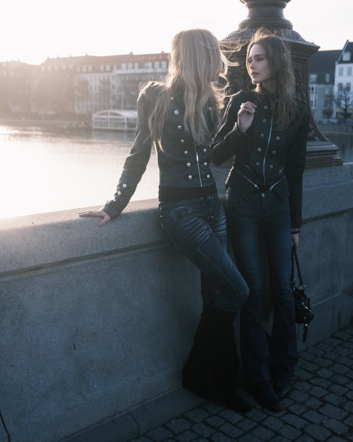 Two women in military-style jackets and dark denim posing by a river at sunset.