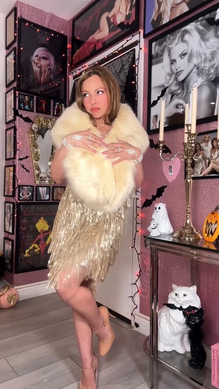 Woman in a gold fringed flapper dress and white fur stole posing in a decorated room.