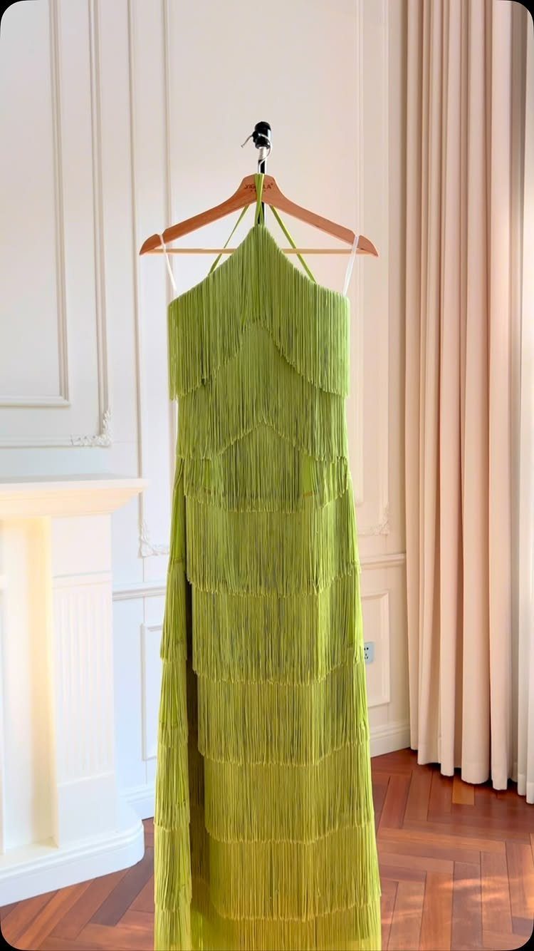 A lime green fringed halter dress hanging on a wooden hanger against a white wall.