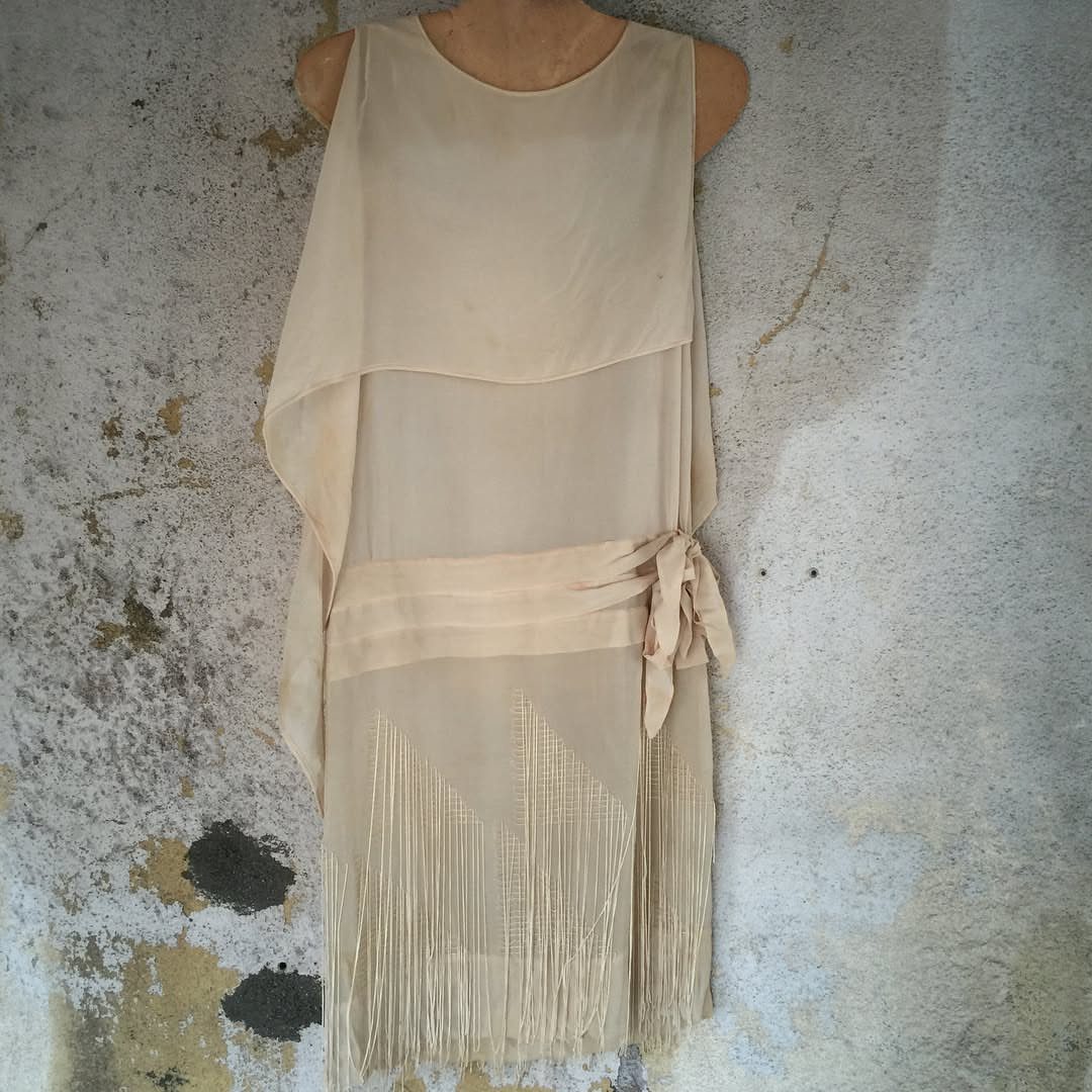 Cream sleeveless dress with long fringe detailing displayed on a mannequin against a textured wall.