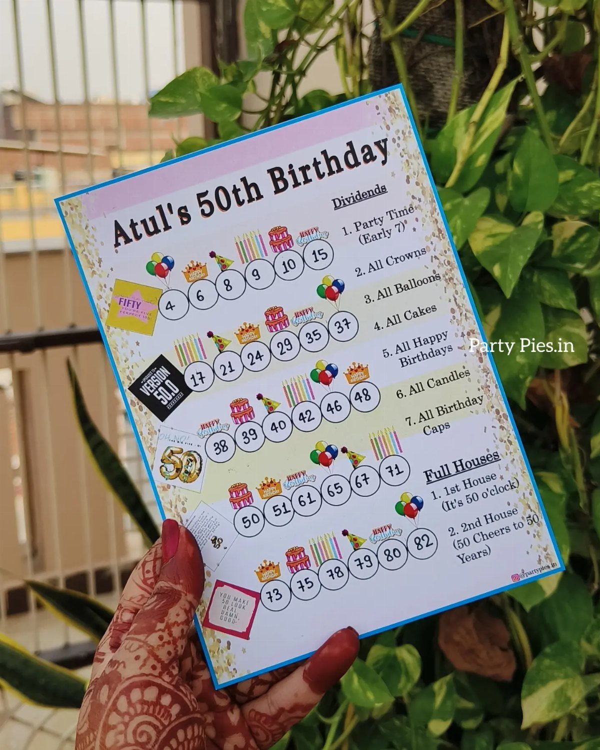 A hand holding a printed bingo-style game card for Atul's 50th birthday celebration.
