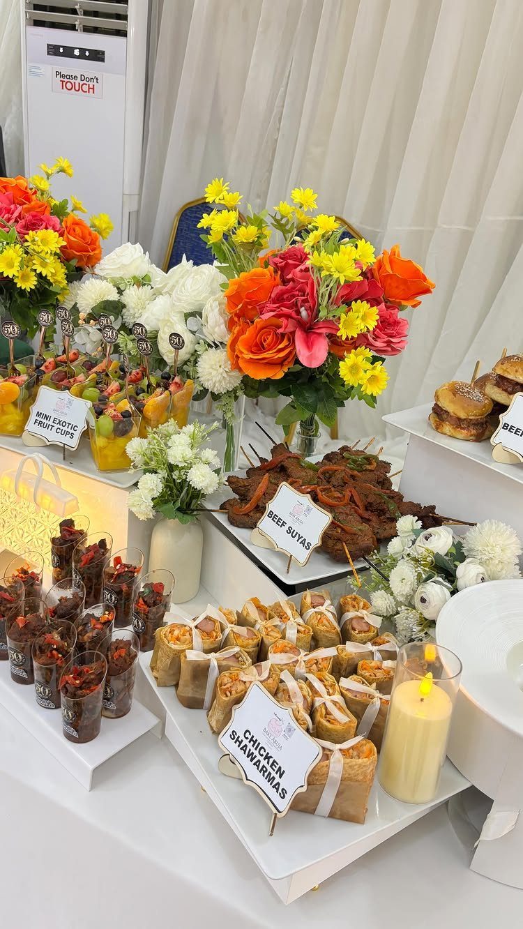 A catered buffet table with various appetizers, colorful flowers, and small food labels on white stands.