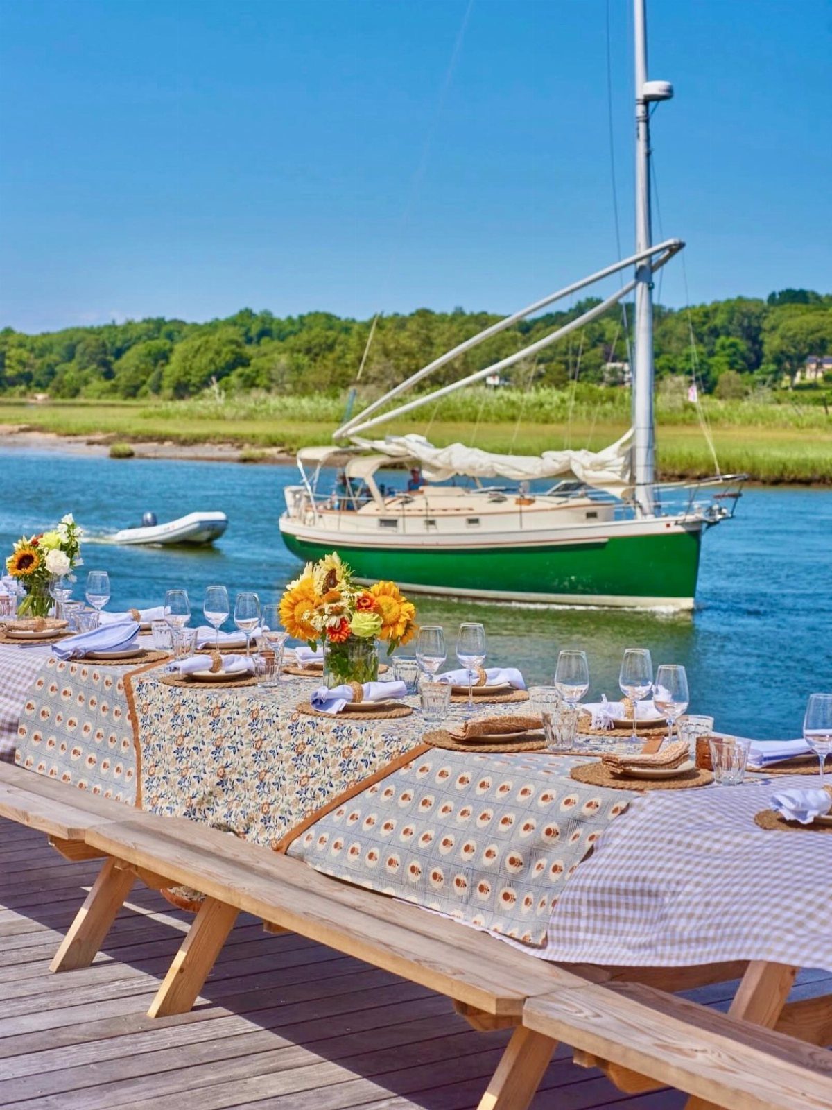 A long outdoor dining table set with floral centerpieces on a wooden pier by a sailboat.