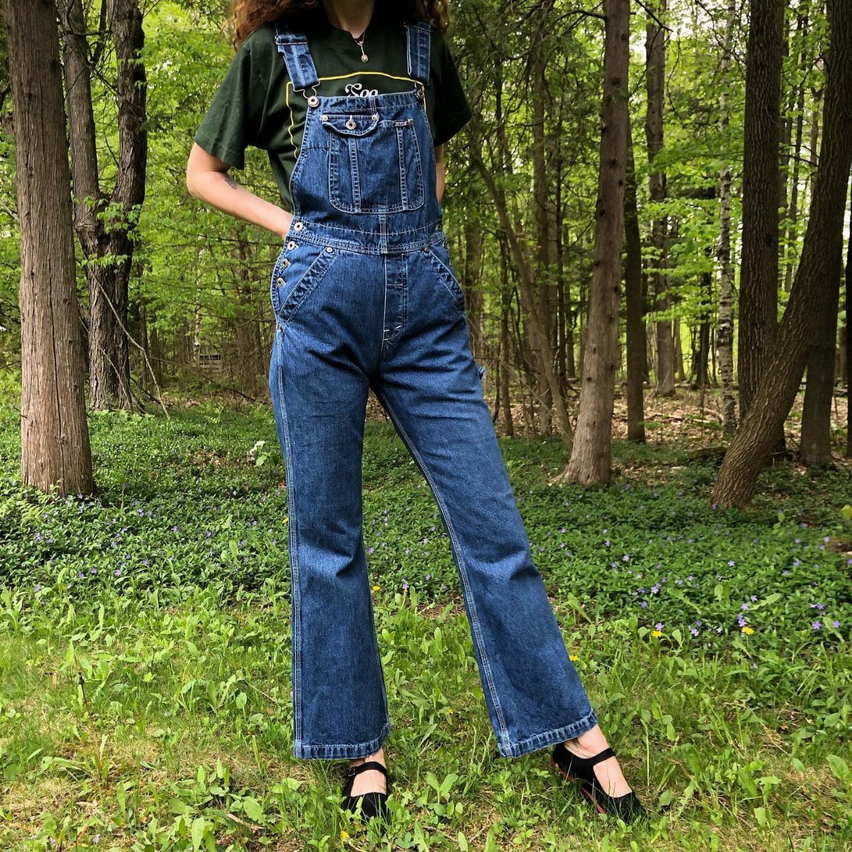 Person wearing dark blue denim overalls and a green t-shirt standing in a forest.