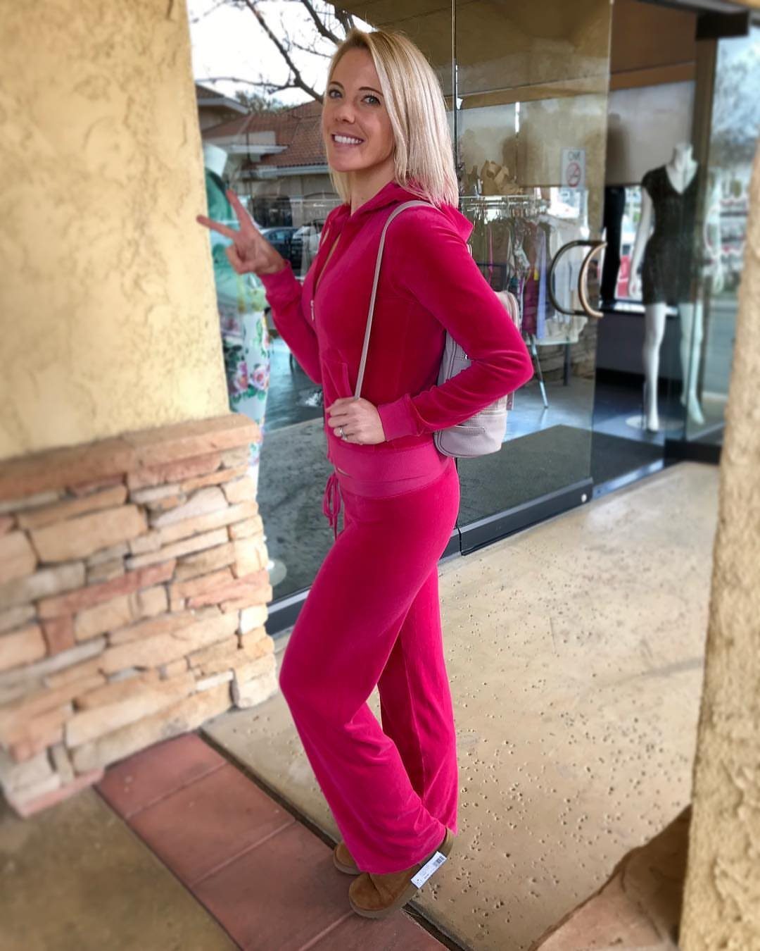 Blonde woman posing in a bright pink velour tracksuit near a glass storefront.