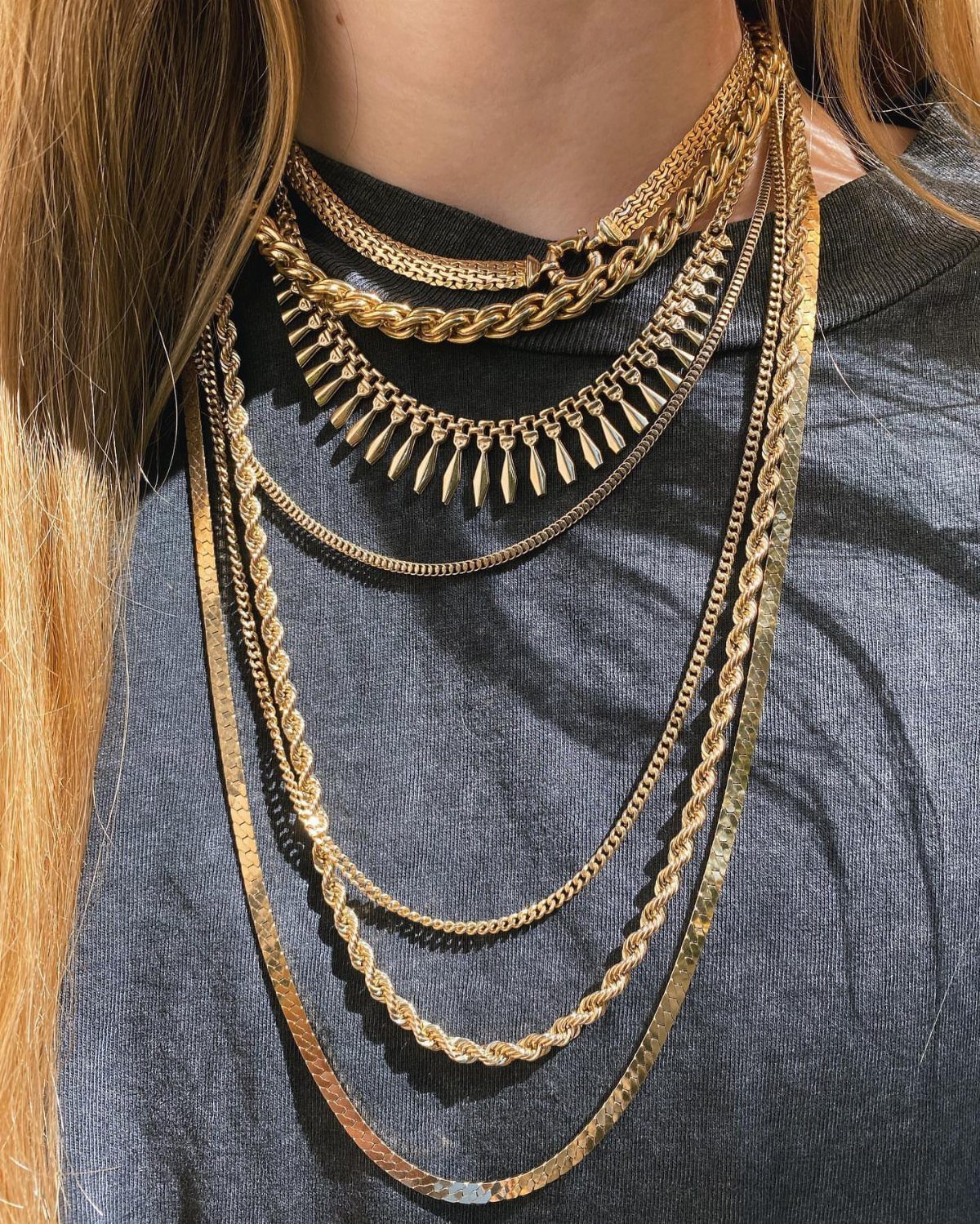 Multiple gold-toned necklaces of different lengths and textures layered over a grey shirt.
