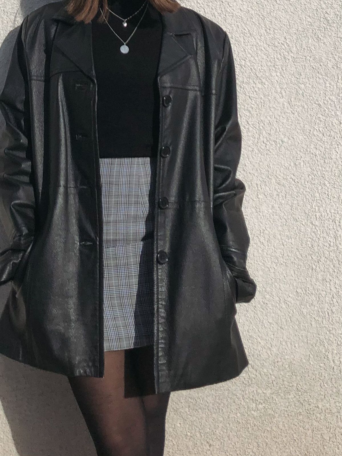 Person wearing an oversized black leather jacket, black turtleneck, and a grey plaid mini skirt.
