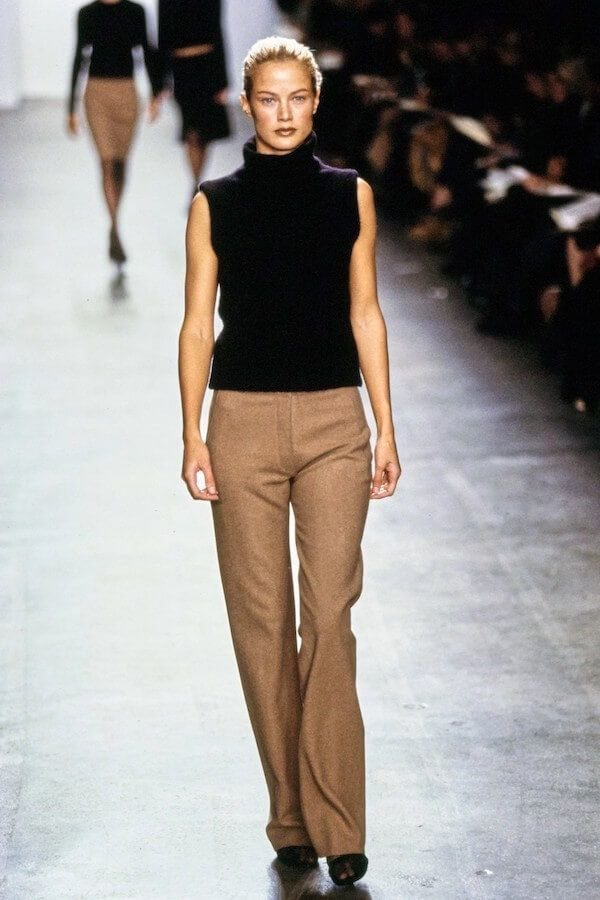 Blonde model walking a runway in a black sleeveless turtleneck and tan flared trousers.