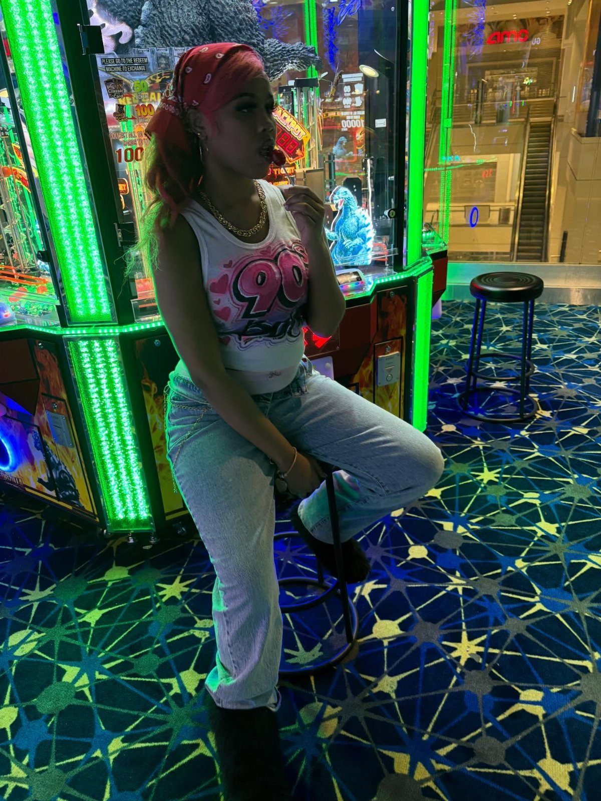 Woman with pink hair wearing a 90s graphic tank top sitting in a neon arcade.