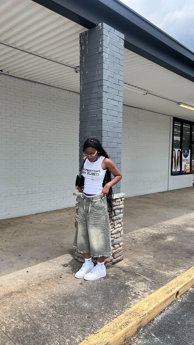 A woman in a white tank top and baggy olive green denim shorts posing by a pillar.