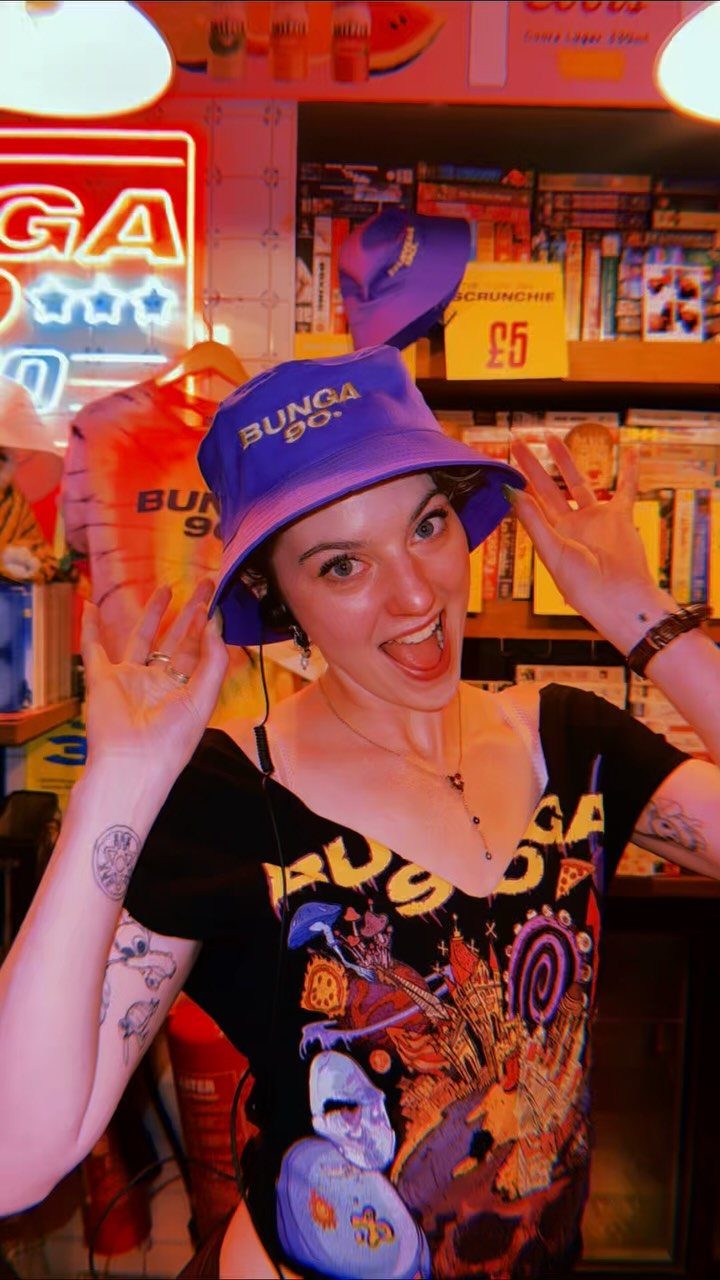Smiling woman wearing a bright purple bucket hat with white text in a colorful shop.