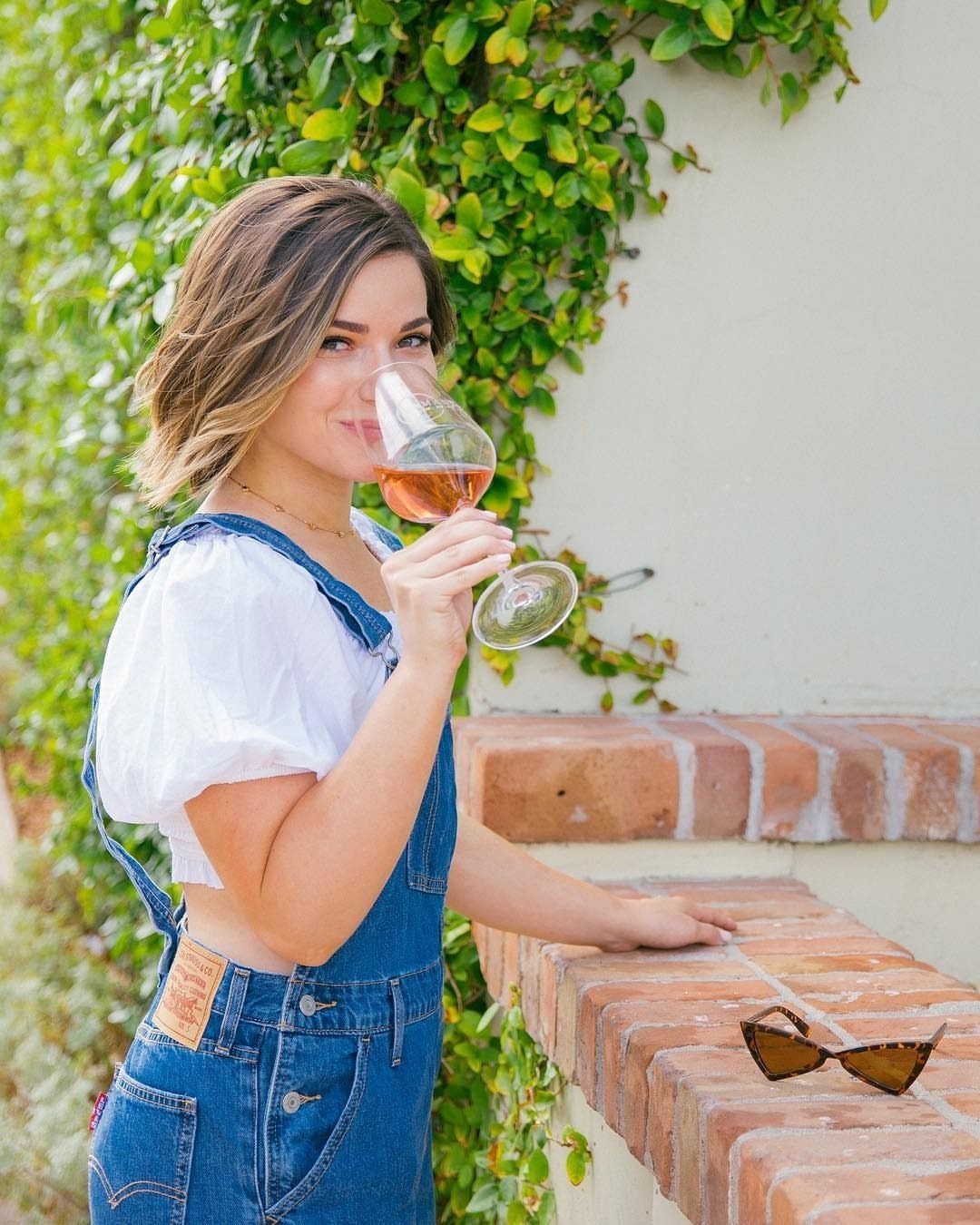 A woman in denim overalls and a white top holding a glass of wine.